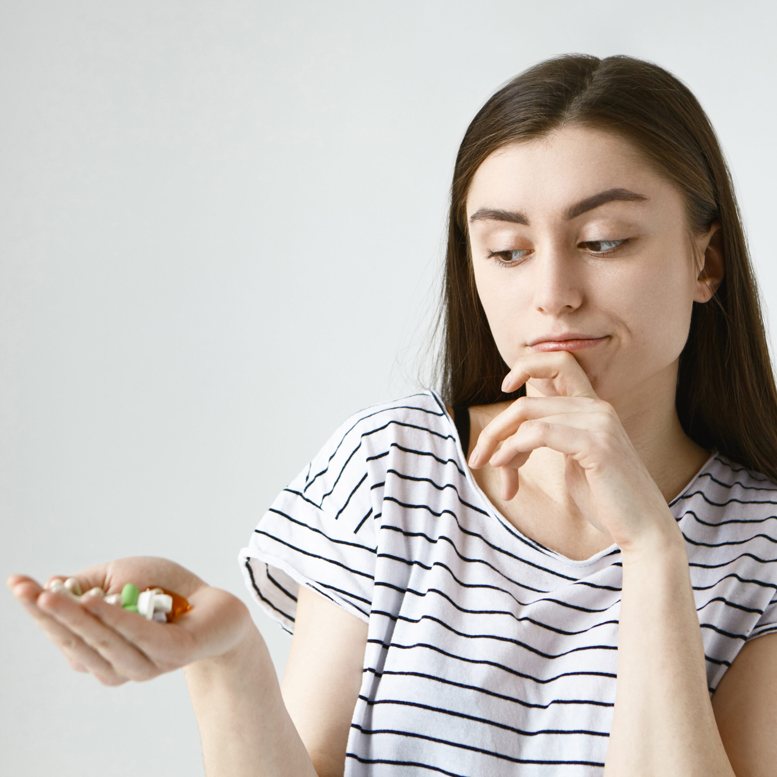 Health, care and treatment concept. Isolated studio shot of indecisive doubtful young lady with loose dark hair rubbing chin thoughtfully and looking at colorful pills on her palm, going to take them